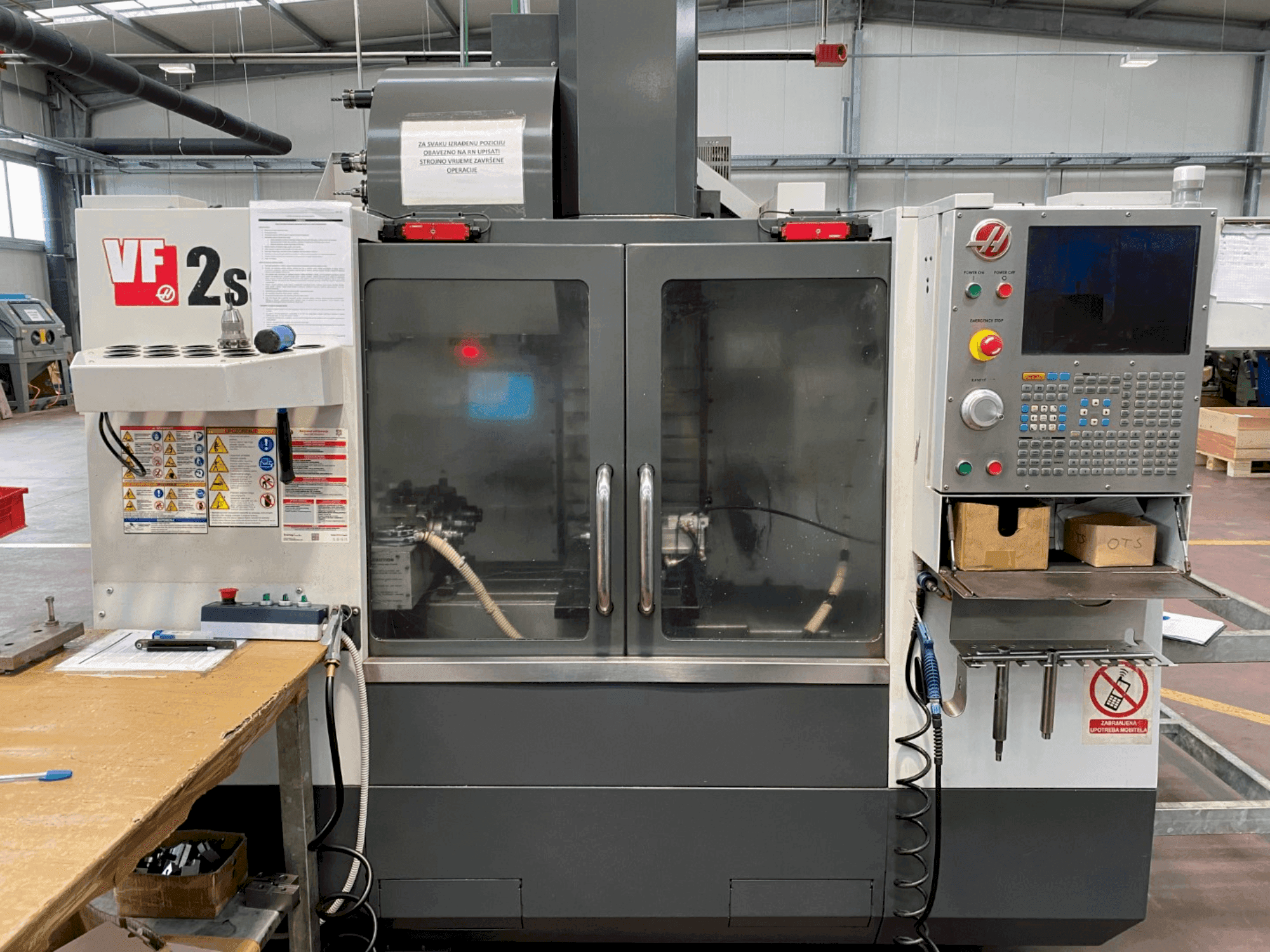 Haas VF-2SS işleme merkezinin önden görünümü, kontrol paneli, soğutma sıvısı hortumu ve yan taraftaki güvenlik etiketleri.